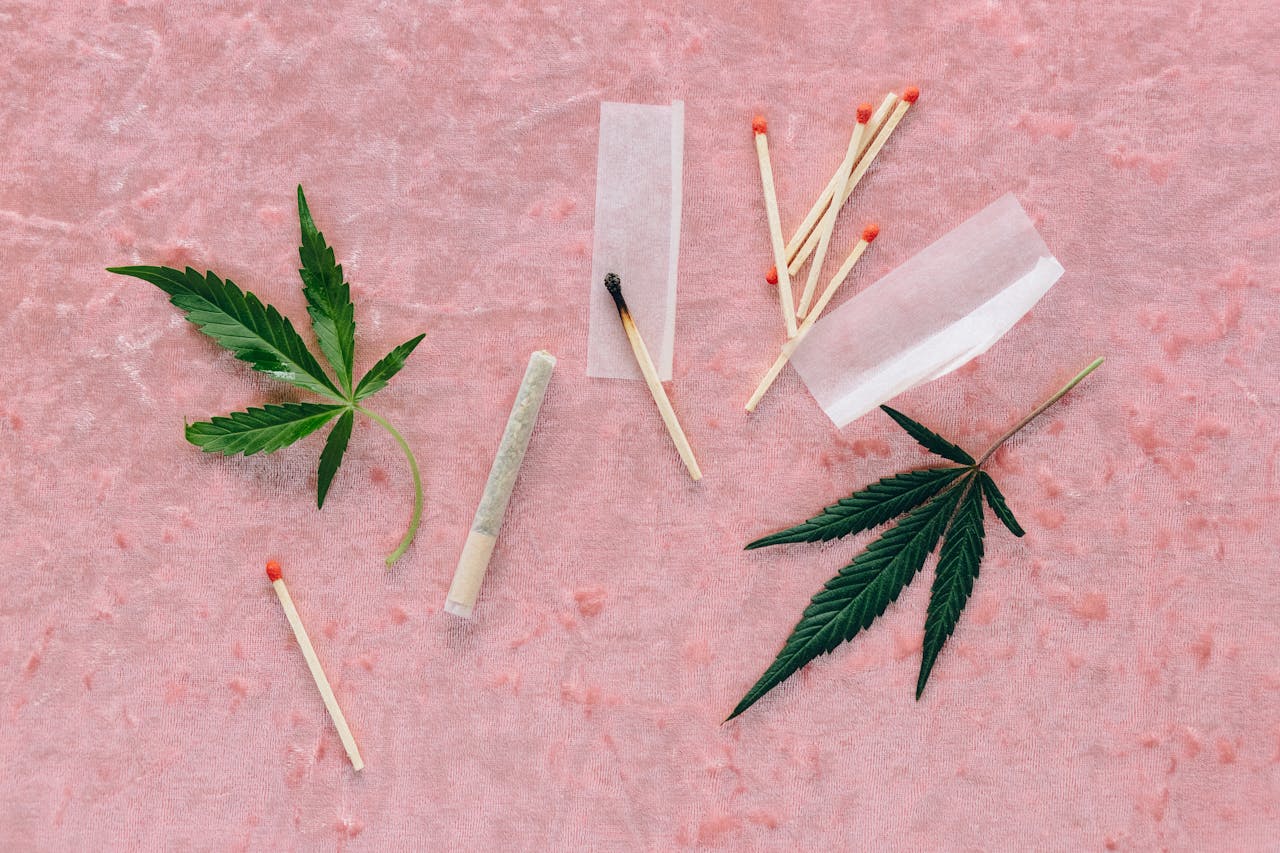 Flat lay of cannabis leaves, rolling paper, and matches on a soft pink background.